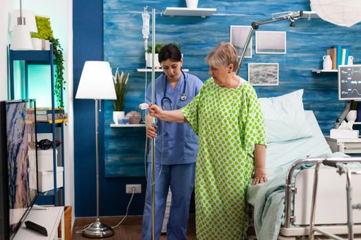 Support cargiver nurse helping retired senior woman caring intravenous drip bag during medicine therapy in living room. Social services nursing elderly retired female. Healthcare assistance