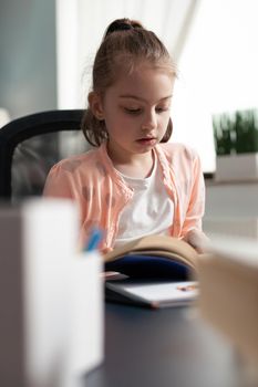 Young kid reading school book for homework deadline while sitting at desk. Little caucasian girl learning lesson from online class notes and preparing for educational knowledge exam