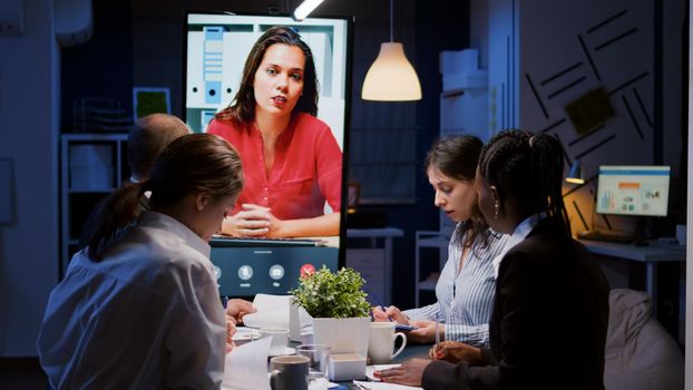 Workaholics overworked multi-ethnic businesspeople discussing with remote manager during online videocall conference meeting late at night in company office room. Focused teamwork working overtime