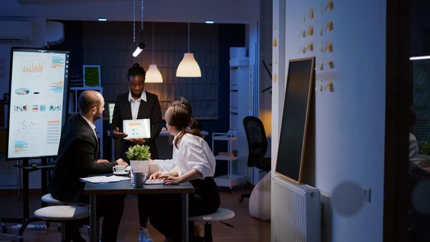 Overworked workaholic businesswoman with dark skin explaining management graphs using tablet overworking in company meeting office room late at night. Multi-ethnic teamwork solving company problem