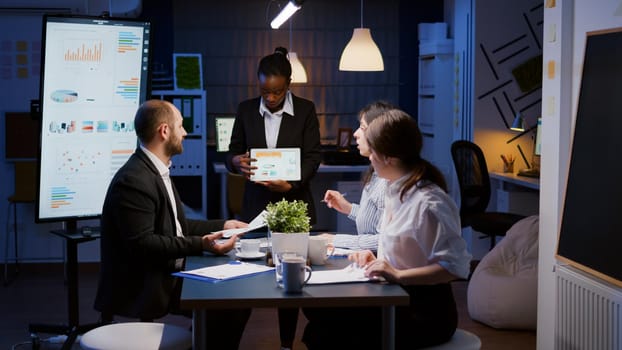 Exhausted workaholic african american showing financial graphs using tablet overworking in company office meeting room. Multi-ethnic businesspeople discussing management statistics late at night