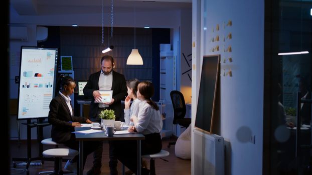 Workaholic overworked businessman explaining company strategy using tablet for presentation working overtime in meeting office room late at night. Diverse multi-ethnic teamwork brainstorming ideas