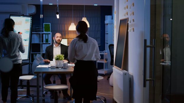 Diverse teamwork overworking in meeting room planning company strategy analyzing management statistics late at night. Multi ethnic coworkers sitting at conference table solving financial presentation