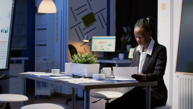 African american businesswoman sitting at conference table drinking coffee while analyzing financial graphs paperwork. Entrepreneur black woman working in office meeting room late at night