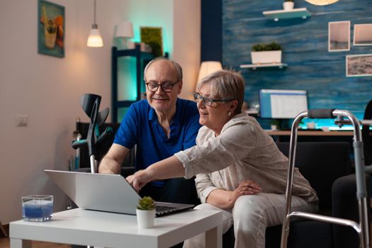 Gray haired woman old man analyzing laptop screen computer monitor at home on sofa. Retired caucasian couple using modern technology device having walk frame crutches and wheelchair