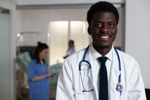 Portrait of african american man working at hospital ward desk with laptop and stethoscope. Black doctor standing, looking at camera with multi ethnic medical staff in background at clinic