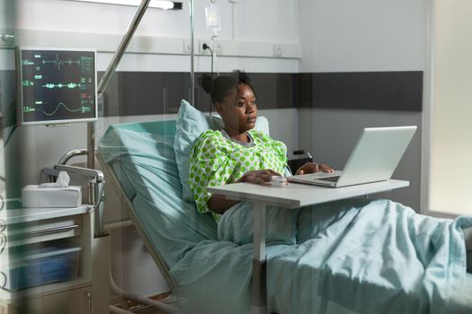 Young woman of african ethnicity browsing internet on laptop in hospital ward bed. African american adult with sickness, disease web surfing on digital device and computer technology