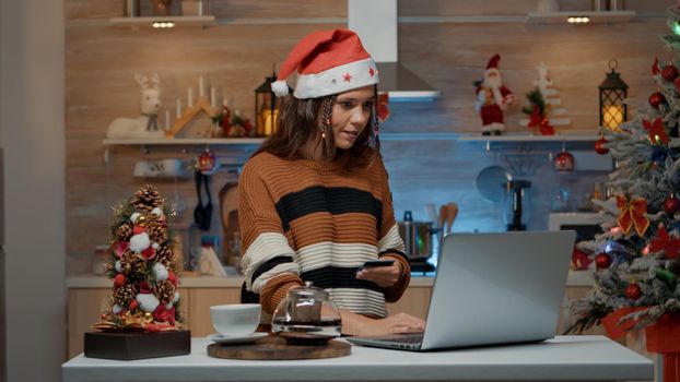 Woman typing credit card information on laptop for buying christmas gifts online. Caucasian young adult doing shopping on internet for festive celebration dinner with friends and family.