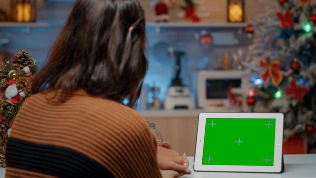 Young woman sitting at home in kitchen with decorations, ornaments and tree looking at green screen background display on tablet. Adult waiting for christmas eve dinner while using device