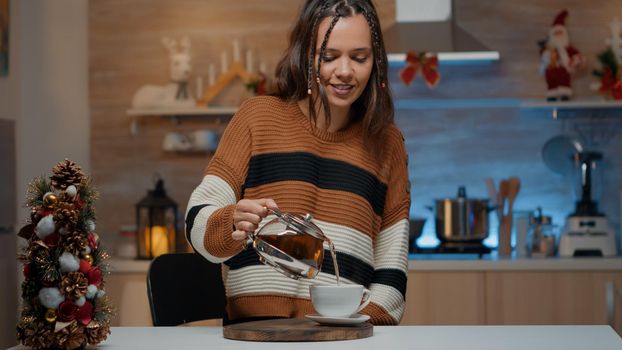 Caucasian woman sitting in christmas decorated kitchen pouring tea from kettle. Young person getting warm with winter beverage while preparing for seasonal holiday celebration party