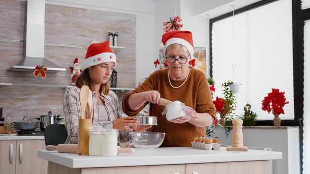 Granddaughter using kitchen strainer while grandmother putting flour ingredient preparing homemade traditional dessert enjoying cooking together. Happy family celebrating christmas holidays