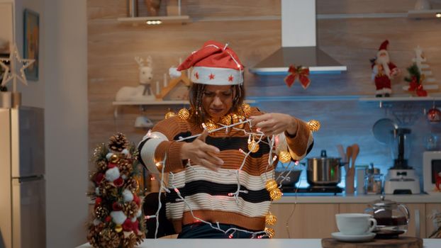 Woman decorating kitchen with festive ornaments while getting tangled in christmas string lights for tree. Young person holding illuminated garland for holiday celebration preparations