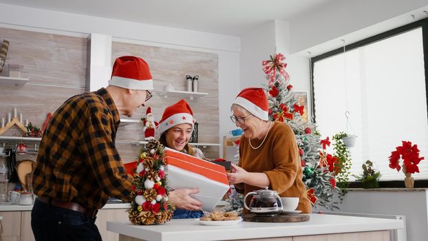 Happy family enjoying winter holiday celebrating christmas season standing at table in decorated kitchen. Grandparents surprising grandchild wrapper present with ribbon on it during christmastime