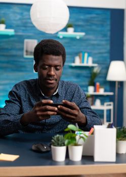 Black african american man using a smartphone at home working on social media platform. Young guy looking at telephone sitting at desk doing online internet search on modern device
