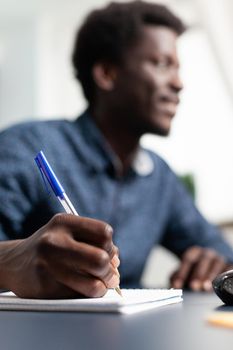 Black business student taking notes on notebook using computer, remote working from home and taking online college classes. African american man learning lessons on internet connection