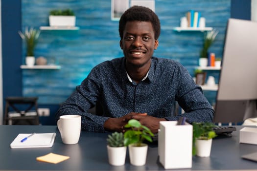 Portrait of african american man smiling into the camera at office desk. Black young man in modern flat apartment working from home using computer technology for digital business