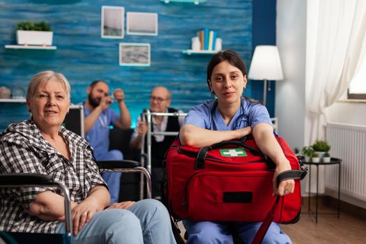 Portrait of support nurse worker holding medicine kit bag sitting beside disabled senior patient looking into camera during medical therapy. Healthcare assistance. Social services nursing at home