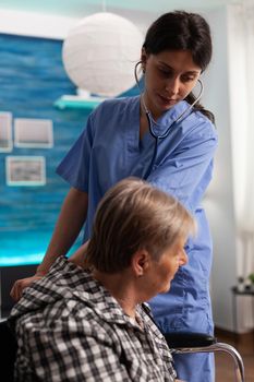 Support nurse worker listening lungs of retired female in wheelchair using medical stethoscope during examination. Social services nursing elderly retired female. Healthcare assistance