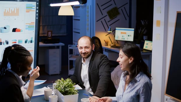 Portrait of businessman looking into camera overworking in company meeting office room late at night. Diverse multi-ethnic workaholics businesspeople analyzing marketing strategy in evening