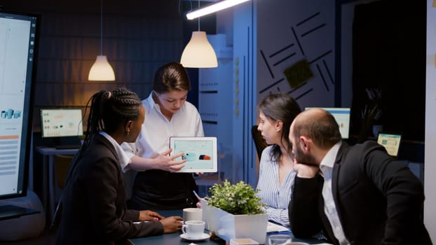 Workaholic focused businesswoman holding tablet explaining company graph overworking in business office meeting room late at night. Diverse multi-ethnic coworkers solving statistics problem in evening