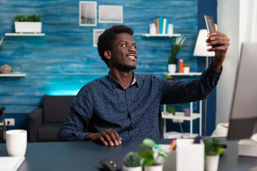 African american man taking selfie in cozy living room while remote working from home. Person using photo for social media profile with mobile smartphone smiling while sitting at desk