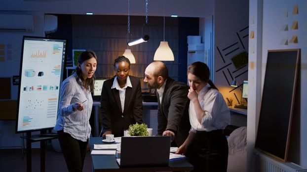 Multi ethnic businesspeople brainstorming company ideas checking graphs statistics using laptop standing at conference table. Diverse teamwork working at management presentation in meeting room