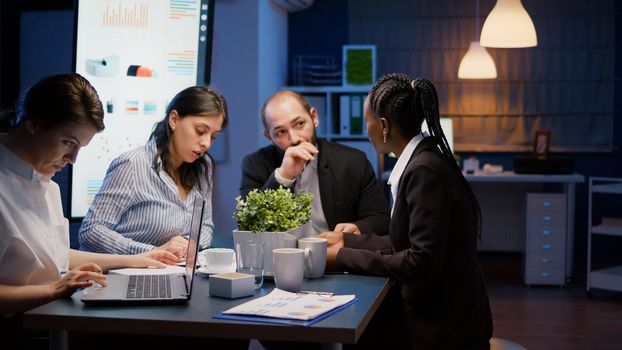 Multi-ethnic businesspeople discussing financial company solution sitting at conference table in meeting room. Diverse teamwork overworking at management strategy using laptop late at night