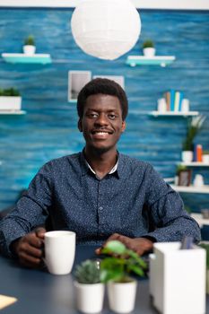 Portrait of african american man smiling into the camera at office desk. Black young man in modern flat apartment working from home using computer technology for digital business