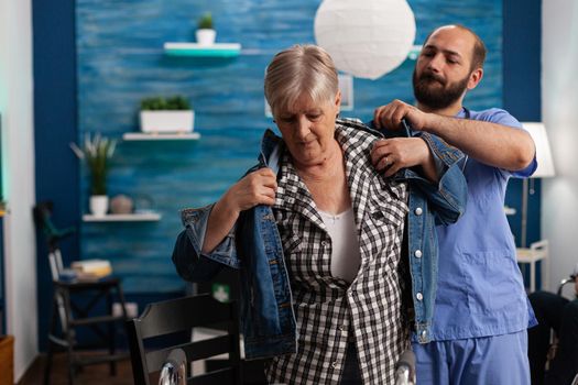 Social asisstant man worker helping pensioner disabled senior woman puts jacket during recovery session. Healthcare assistance. Social services nursing elderly retired woman. Disability people support