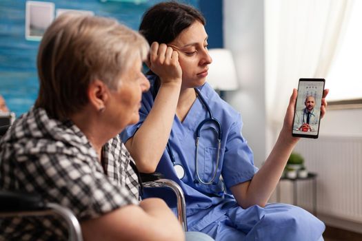 Social worker holding smartphone for disabled senior retired woman in wheelchair during online medical video call teleconference with physician doctor. Healthcare treatment in nursing home