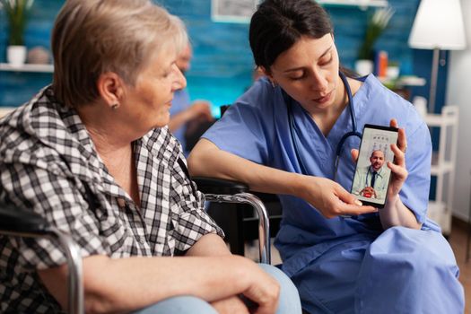 Social worker holding smartphone for disabled senior retired woman in wheelchair during online medical video call teleconference with physician doctor. Healthcare treatment in nursing home