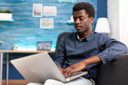 African american man typing on modern laptop computer relaxing on couch in living room. Black person remote working from home, using connection social media online internet for job