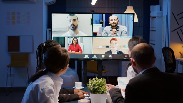 Multi ethnic businesspeople discussing company statistics with remote coworkers during online videocall conference meeting brainstorming ideas. Diverse teamwork working overtime in corporate room