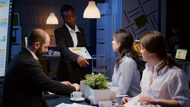 Entrepreneur woman with dark skin overworking in company office meeting room explaining management strategy using tablet. Workaholics multi-ethnic businesspeople brainstorming late at night