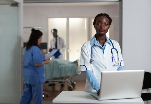 Portrait of african american woman with doctor occupation in hospital ward. Black surgeon, medic looking at camera with stethoscope, multi ethnic working team and patient at clinic