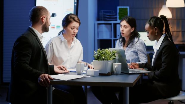 Multi ethnic diverse business teamwork brainstorming management ideas working at company presentation in office meeting room late at night. Black leader writing corporate statistics on laptop