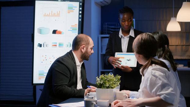 Focused african american businesswoman showing company graphs on tablet working in office meeting room late at night. Diverse multi-ethnic teamwork brainstorming corporate strategy ideas