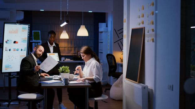 Focused workaholic entrepreneur woman with dark skin explaining management strategy using tablet. Business diverse multi-ethnic teamwork overworking in company office meeting room late at night