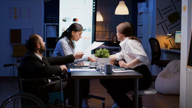 Overworked paralyzed disabled entrepreneur man in wheelchair sitting at desk overworking in company office meeting room. Manager is ignored while coworkers analyzing statistics paperwork late at night