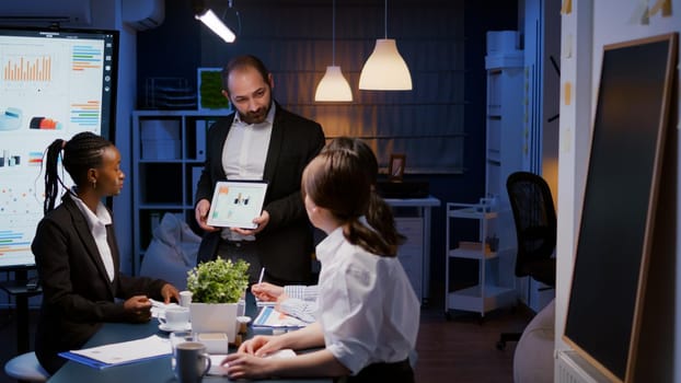 Overworked businessman showing financial graphs presentation using tablet brainstorming company ideas. Workaholic diverse multi-ethnic businesspeople overworking late at night in office meeting room