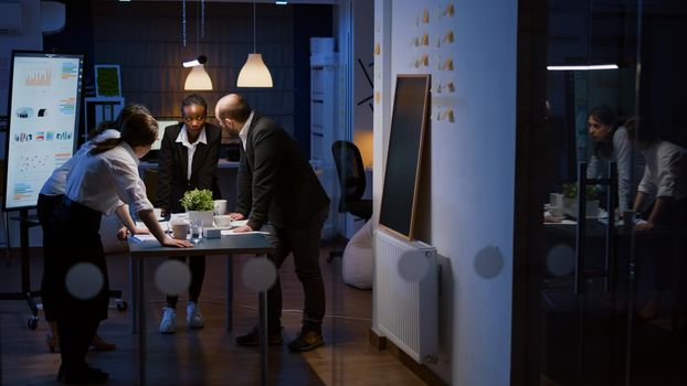 Diverse multi ethnic businesspeople teamwork lean on conference table having brainstorming session discussing company presentation. Coworkers overworking in meeting room late at night