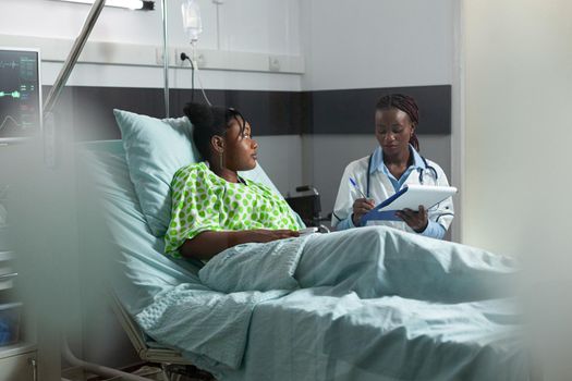 African american young adult with disease talking to doctor while sitting in hospital ward. Black surgeon listening to ill patient in bed, helping with professional advice and medicine