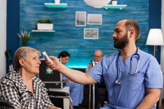 Assistant man helper checking temperature using medical infrared thermometer discussing with senior woman during therapy. Social services nursing elderly retired female. Healthcare assistance