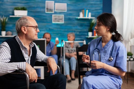 Support assistant worker explaining healthcare treatment to disabled senior man in wheelchair during medical consultation in living room, Social services nursing elderly retired male