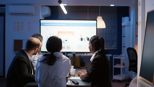 Businesswoman explaining financial graphs using presentation monitor overworking in office meeting room in evening. Diverse multi-ethnic teamwork brainstorming company strategy at night