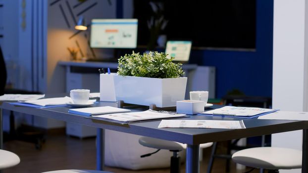 Interior of professional empty start-up business office meeting room with nobody in it. On table standing financial documents late at night. In background computer with company graph on monitor