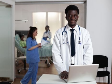 Portrait of african american man working at hospital ward desk with laptop and stethoscope. Black doctor standing, looking at camera with multi ethnic medical staff in background at clinic