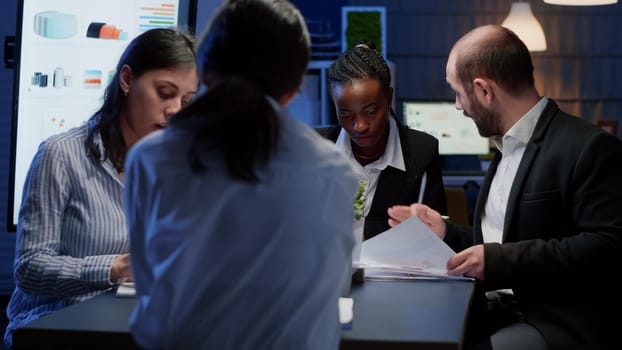 Diverse business entrepreneurs sitting at conference table working at management solution presentation in office meeting room late at night. Multi ethnic teamwork brainstorming ideas