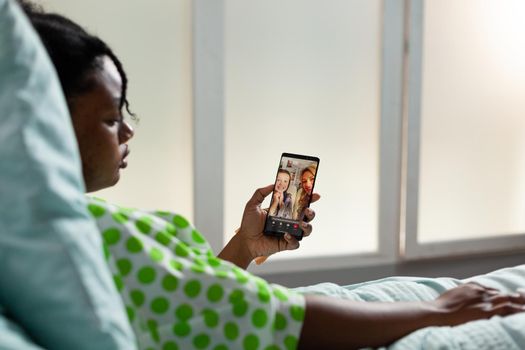 Patient of african american ethnicity using smartphone for video call with family. Young woman laying in hospital ward bed talking to people on online internet conference while recovering