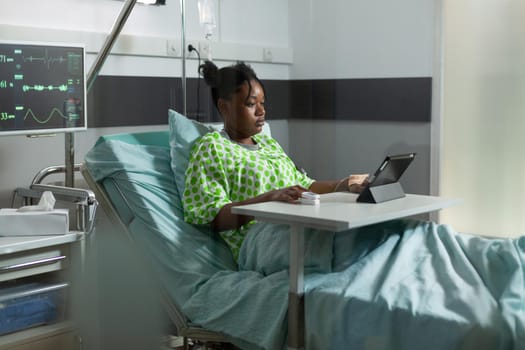 Young african american woman using tablet in hospital ward bed. Sick patient with online technology recovering from disease at clinic with medical equipment. Person web surfing